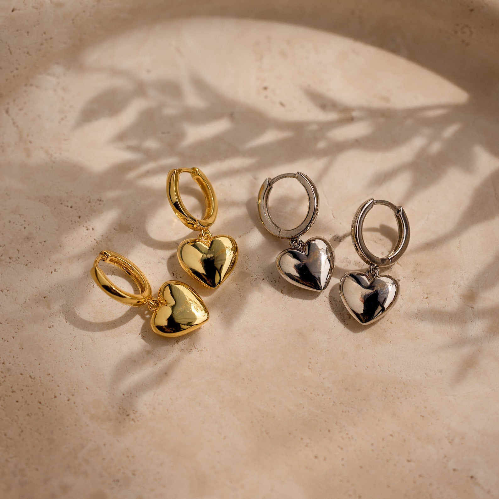 A pair of Puff Heart Charm Huggies in 18K Gold with a dainty heart-shaped design, displayed on a beige textured surface.