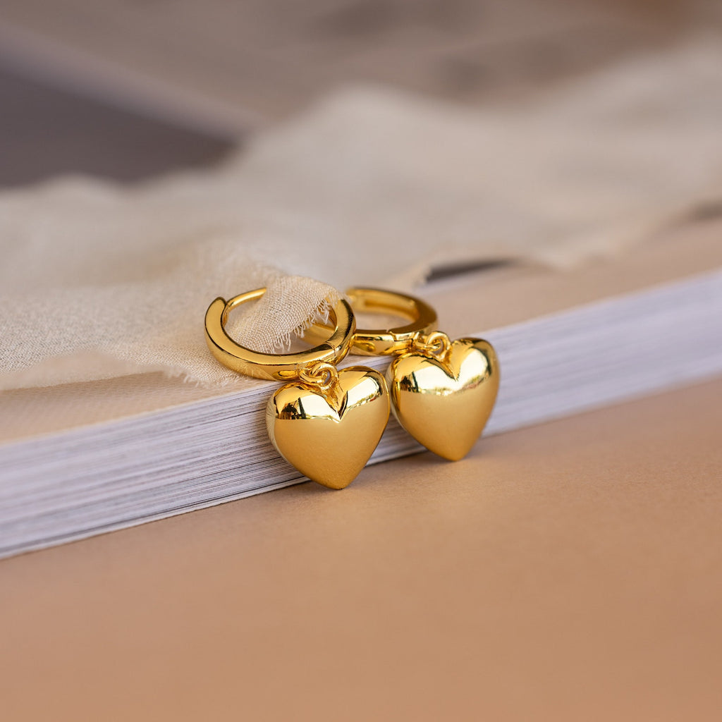 Two Puff Heart Charm Huggies in 18K Gold rest on the edge of a closed book.