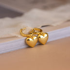 Two Puff Heart Charm Huggies in 18K Gold rest on the edge of a closed book.