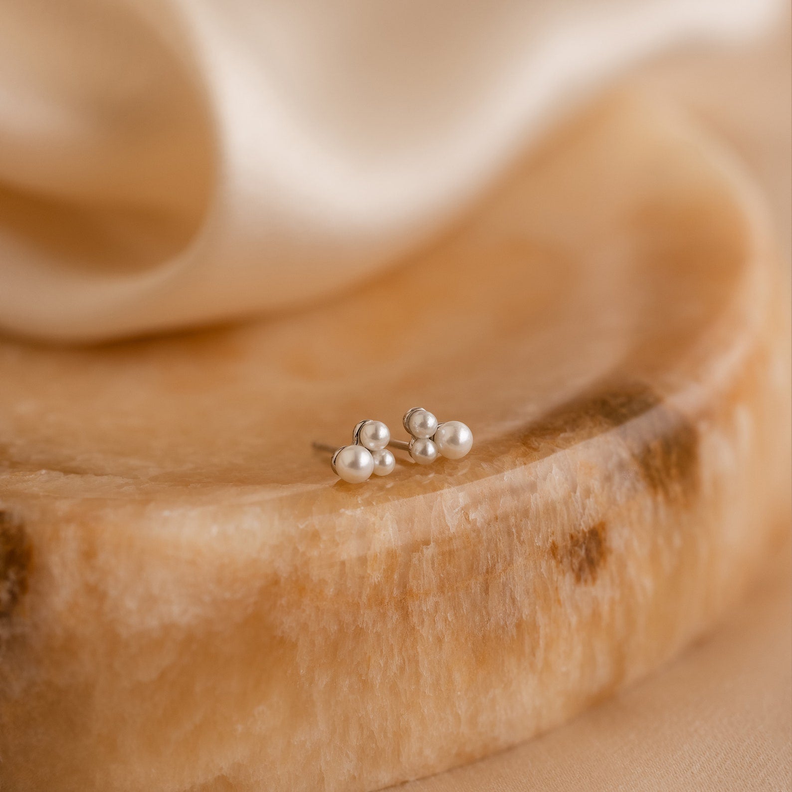 The Trio Cluster Pearl Studs are displayed on a polished stone surface with a soft beige fabric background—ideal wedding jewelry for an elegant bridal look.