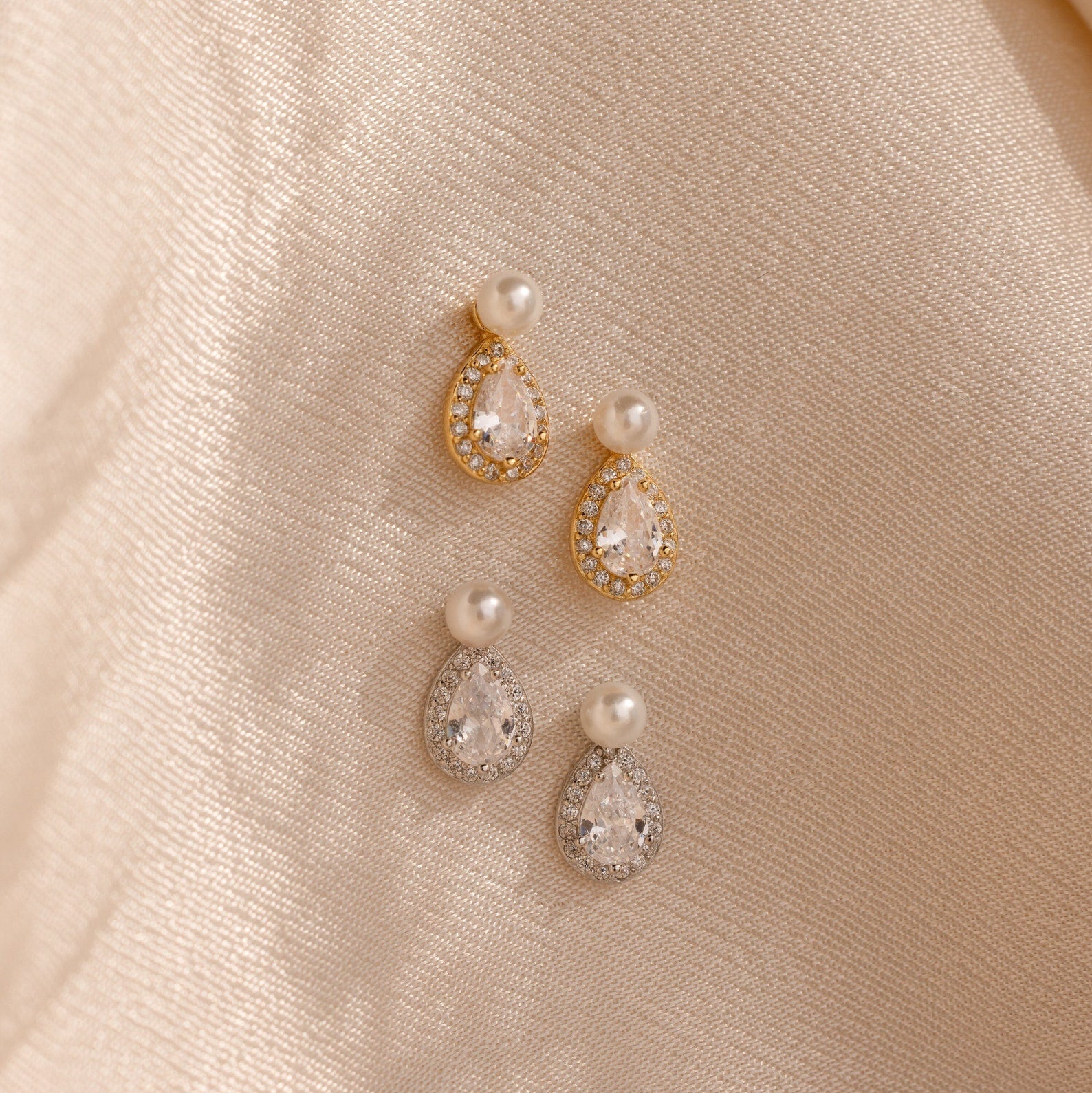 Four pairs of Pearl Diamond Teardrop Studs are elegantly displayed on a soft, light beige fabric background.