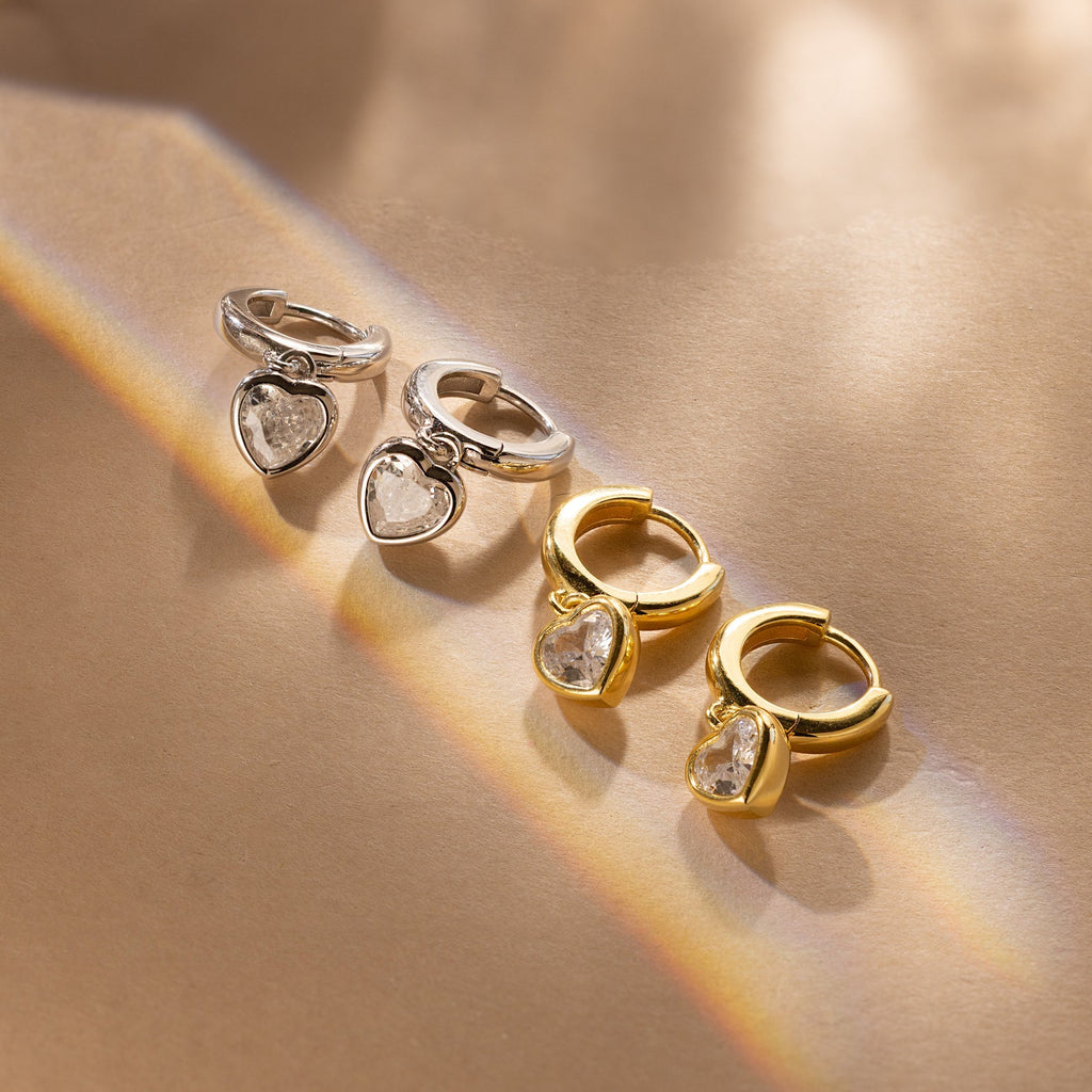 Two pairs of Diamond Heart Drop Huggies—one in silver and one in gold—each features a dangling coquette-inspired heart charm for a playful yet elegant touch.