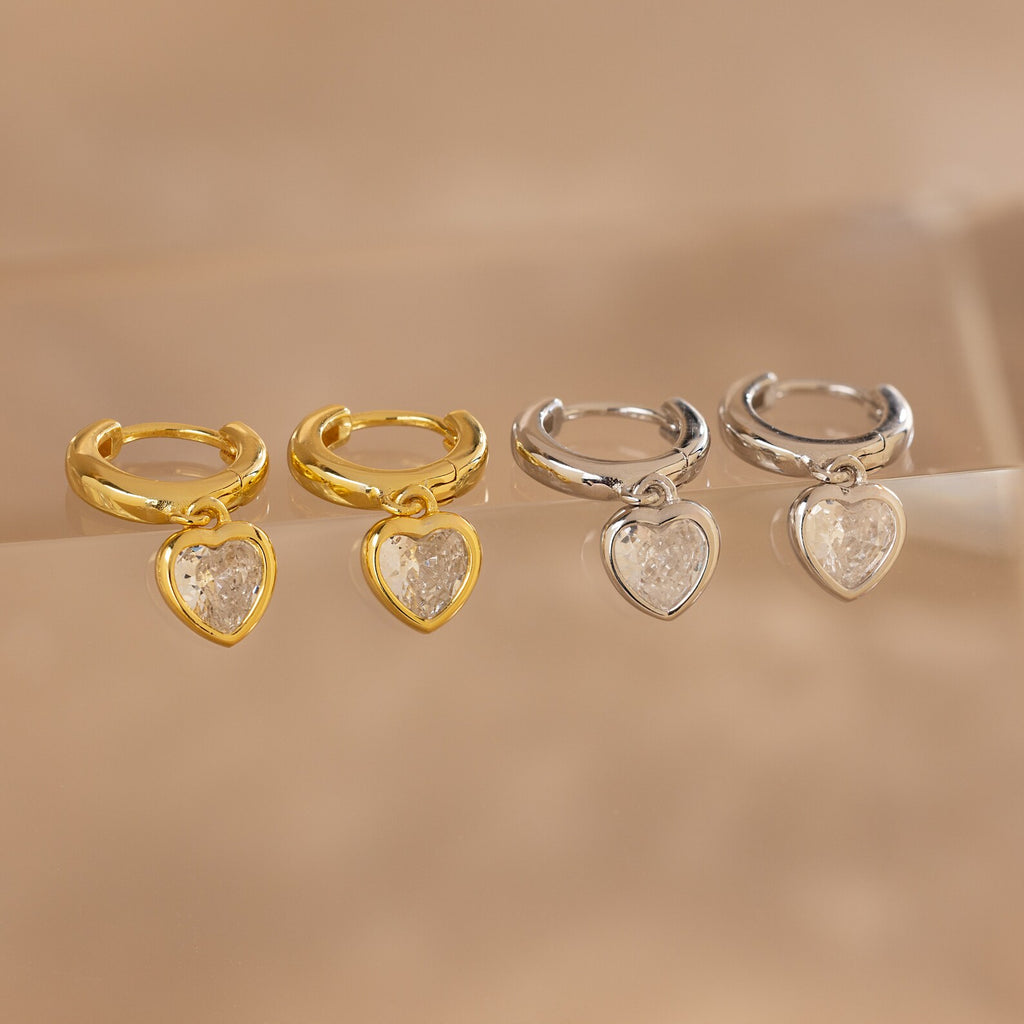 Two pairs of Diamond Heart Drop Huggies—coquette-inspired heart-shaped earrings in gold and silver, each with a clear gemstone at the center—add charming sparkle to any look.