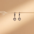 Diamond Heart Drop Huggies, silver hoop earrings with coquette-inspired heart-shaped pendants, hang from a horizontal straw on a beige background, showcasing playful elegance.