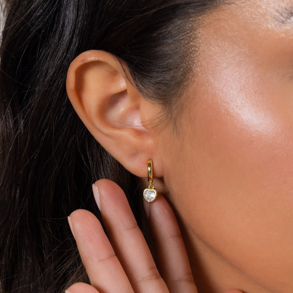 Close-up of a woman's ear wearing the Diamond Heart Drop Huggies, a gold hoop earring with a diamond heart charm pendant in coquette-inspired style, as her hand rests near her face.