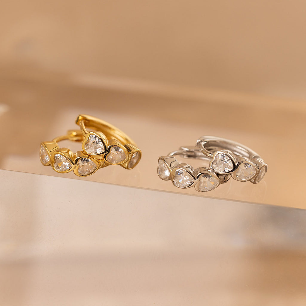Two rings with heart-shaped stones—one gold, one silver—are displayed on a clear surface, pairing perfectly with the Diamond Bezel Heart Huggies.