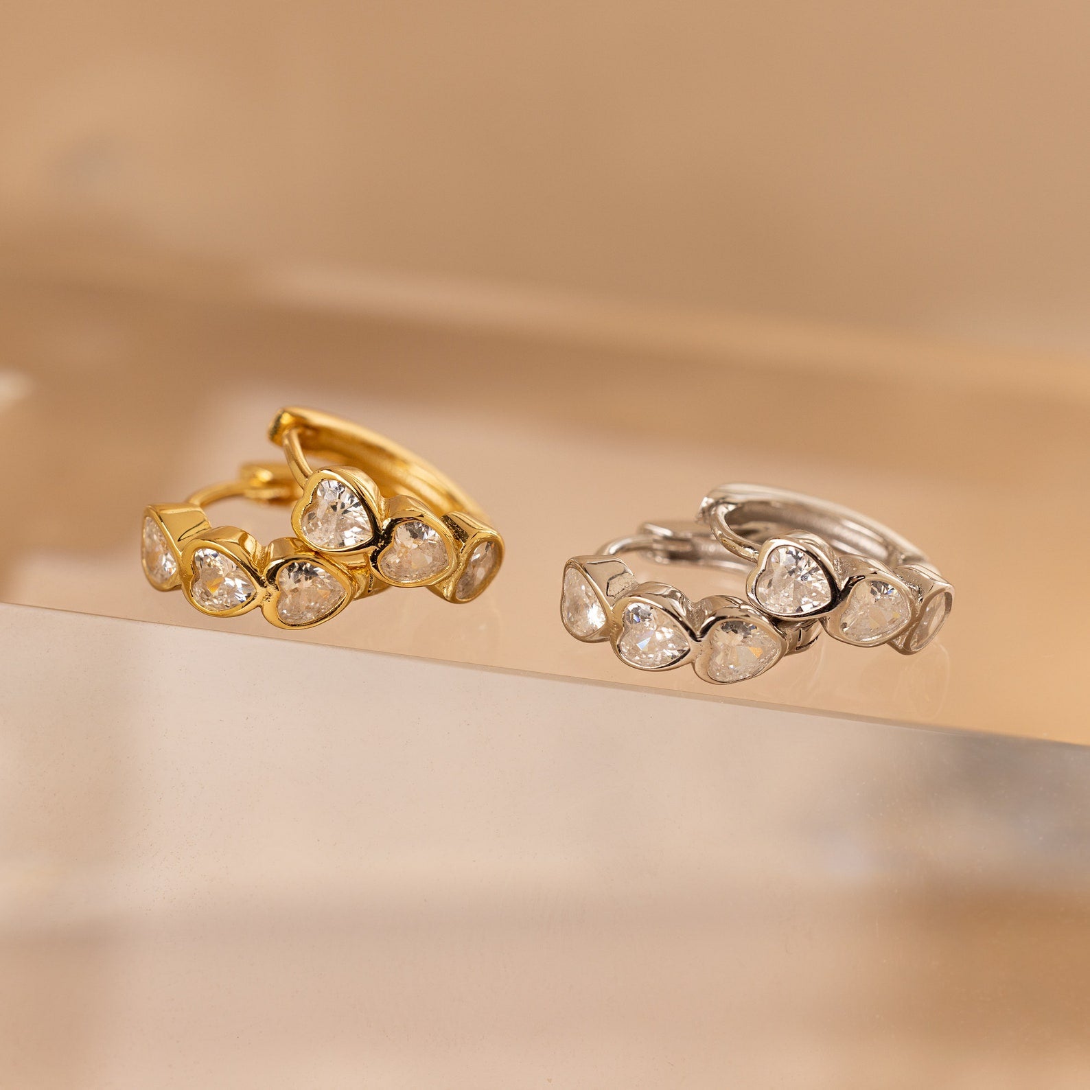 Two rings with heart-shaped stones—one gold, one silver—are displayed on a clear surface, pairing perfectly with the Diamond Bezel Heart Huggies.