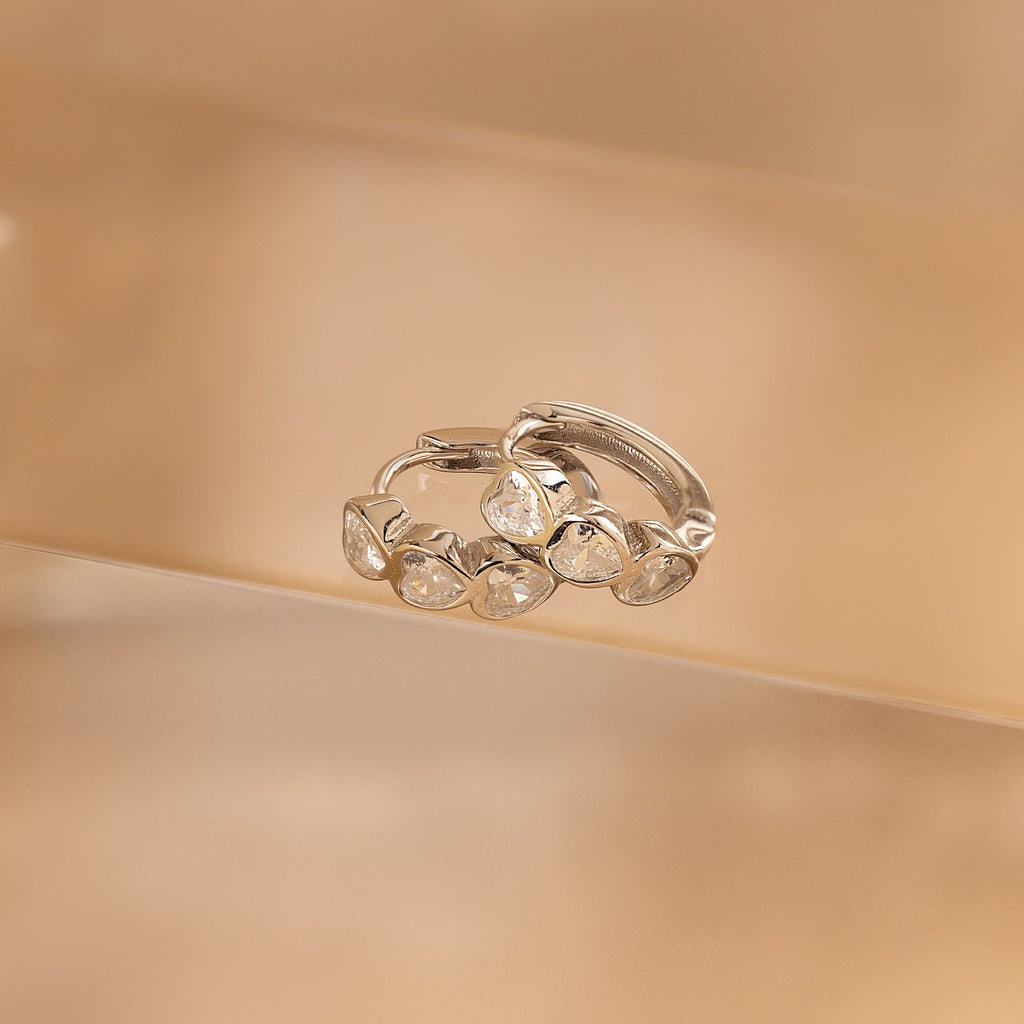 Diamond Bezel Heart Huggies featuring five sparkling gemstones each are showcased against a neutral beige background.