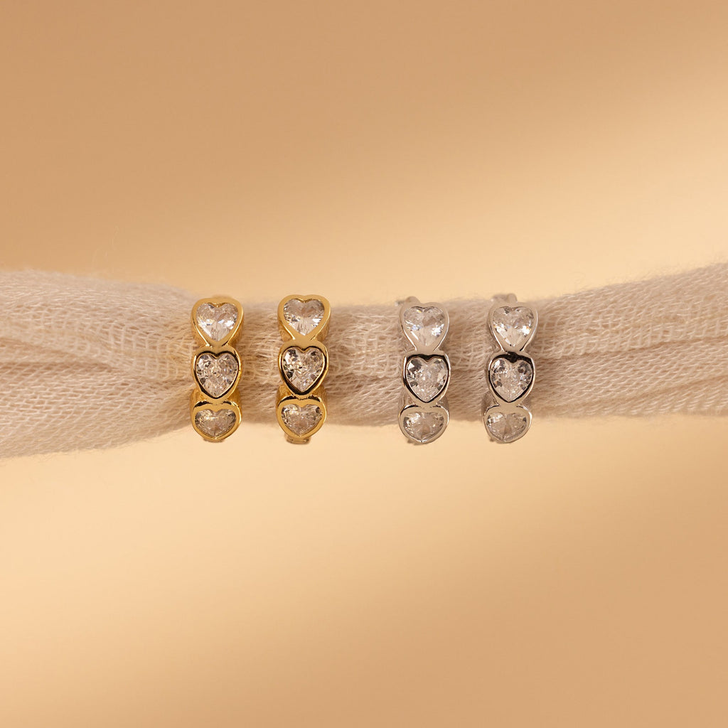 Four heart-shaped gemstone rings in gold and silver rest on beige fabric, beautifully complemented by our Diamond Bezel Heart Huggies for a romantic touch.