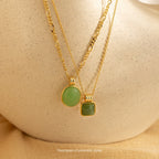 Two sophisticated Mystery Necklaces with green stone pendants rest on a textured beige surface, adding a touch of intrigue to any jewelry collection.