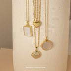 Four Mystery Necklaces, featuring heart and round gold lockets, are displayed on a textured cream background—elegant pieces that add a touch of intrigue to any jewelry collection.