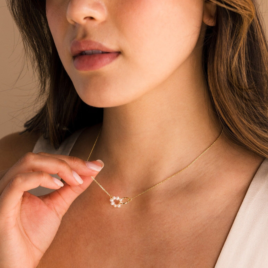 A woman gently touches a delicate gold necklace with a small floral pendant, elegantly layered with the Interlocking Pearl Necklace for added charm.