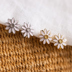 Two pairs of Daisy Huggies—one in silver and one in gold—are displayed on fabric and woven material, offering minimalist flower earrings ideal for jewelry lovers.