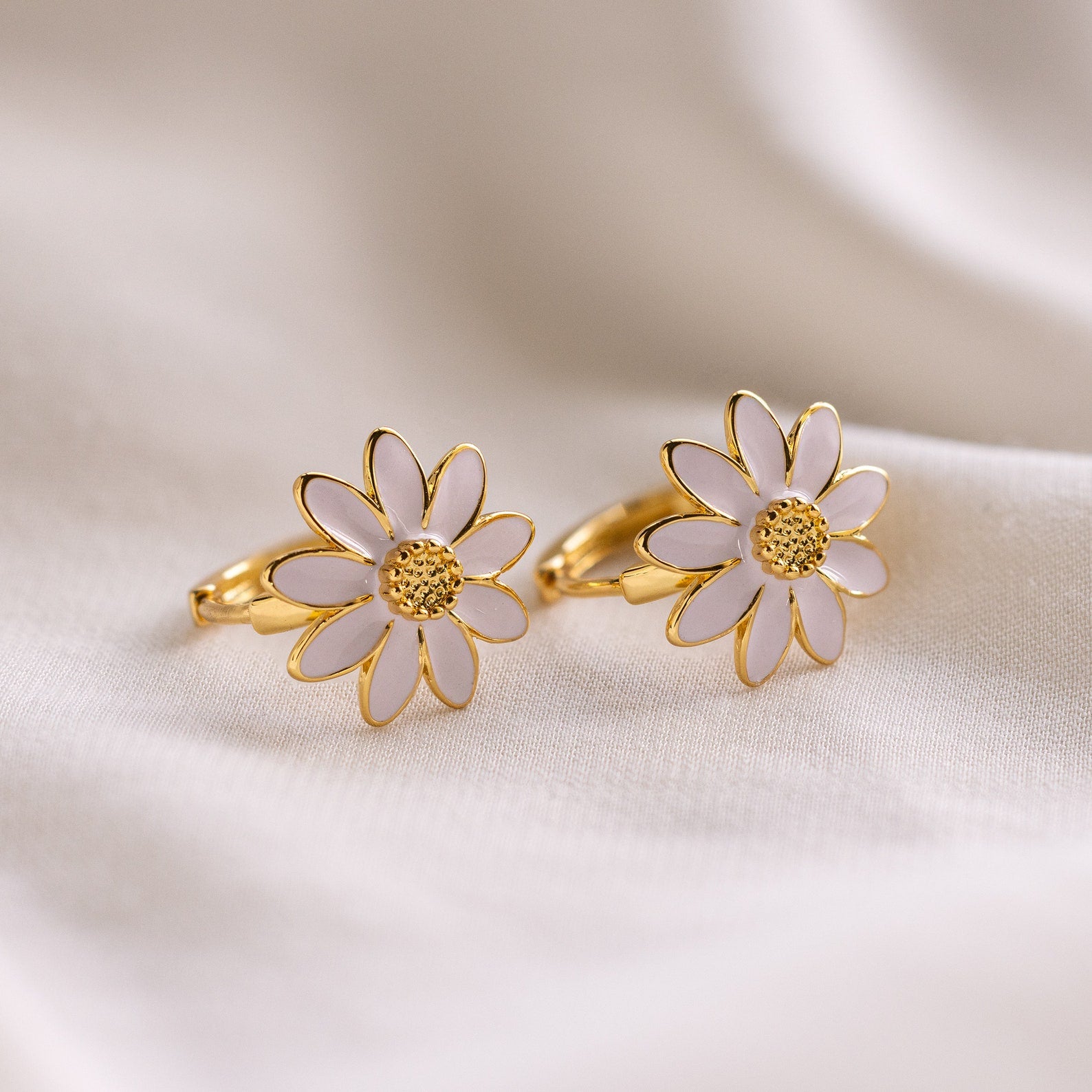 Daisy Huggies: minimalist gold rings featuring white daisy flower designs, ideal for subtle elegance, displayed on a soft, cream fabric backdrop.
