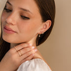 A woman with long brown hair smiles softly, gently touching her neck while wearing Marquise Diamond Flower Studs and rings.