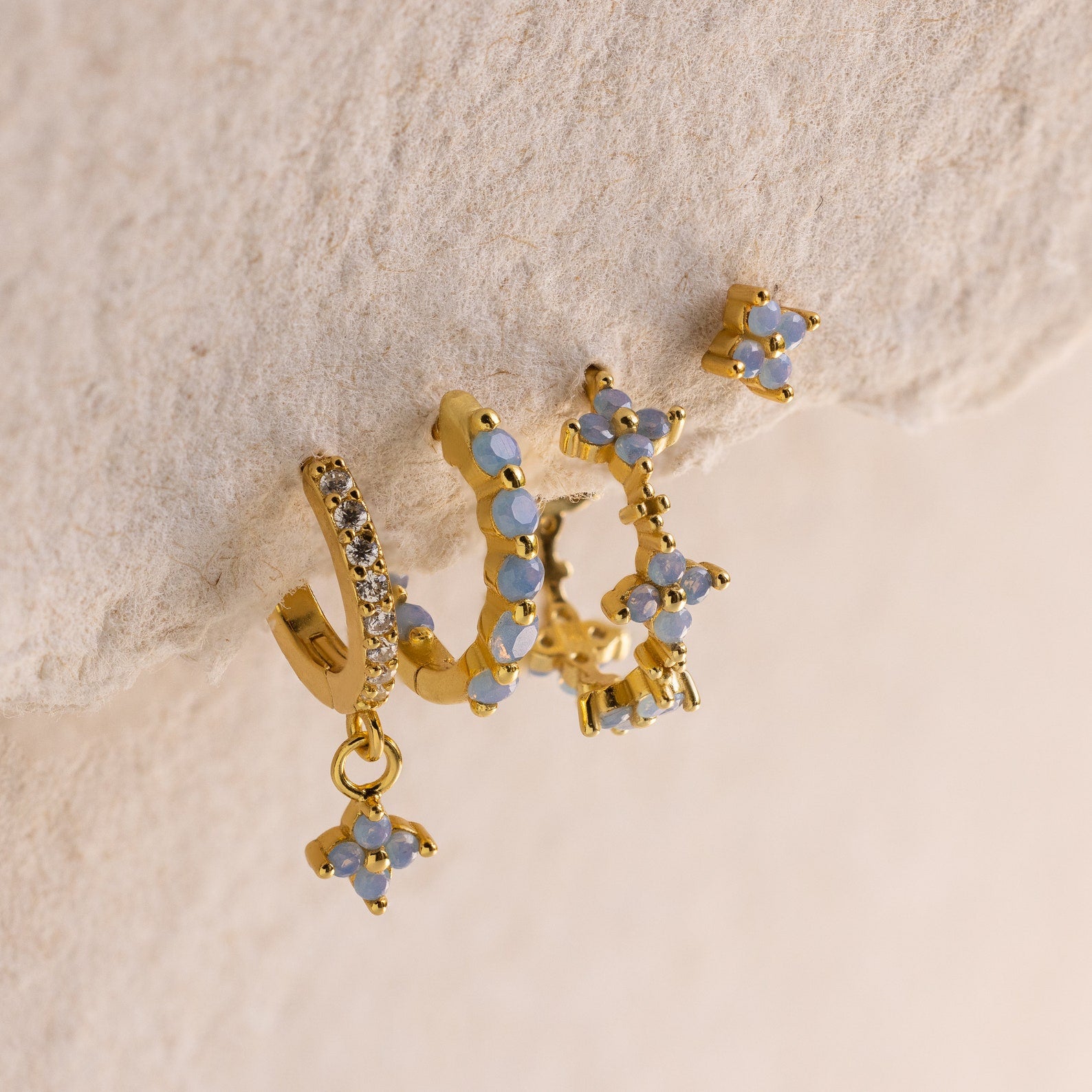 Four gold Inside Out Ivy Huggies, featuring small blue flower details and light blue gemstones, are displayed on a textured beige surface.