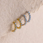 Three hoop earrings—one gold and two silver—featuring light blue gemstones are displayed on a textured beige surface, highlighting the delicate Inside Out Ivy Huggies design.