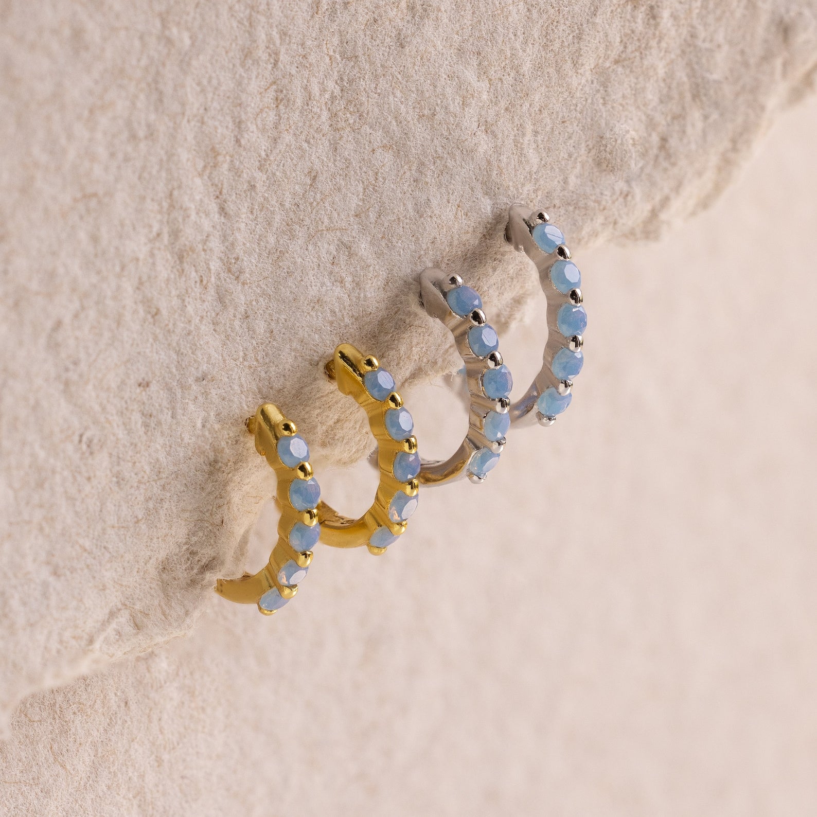 Three hoop earrings—one gold and two silver—featuring light blue gemstones are displayed on a textured beige surface, highlighting the delicate Inside Out Ivy Huggies design.