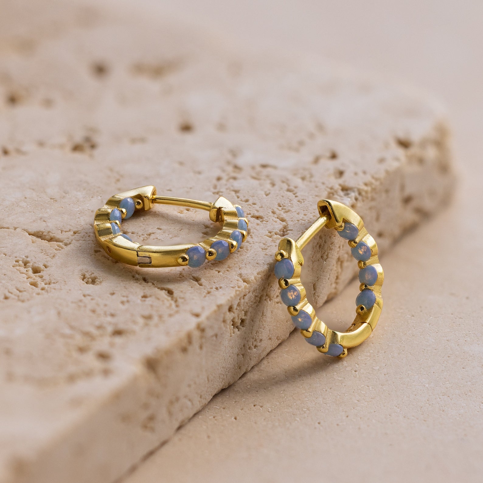 Inside Out Ivy Huggies featuring light blue gemstones are displayed on a textured beige stone surface.