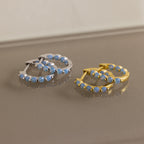 Two pairs of Inside Out Ivy Huggies—one in silver, one in gold—each set with delicate light blue gemstones, are elegantly displayed on a reflective surface.