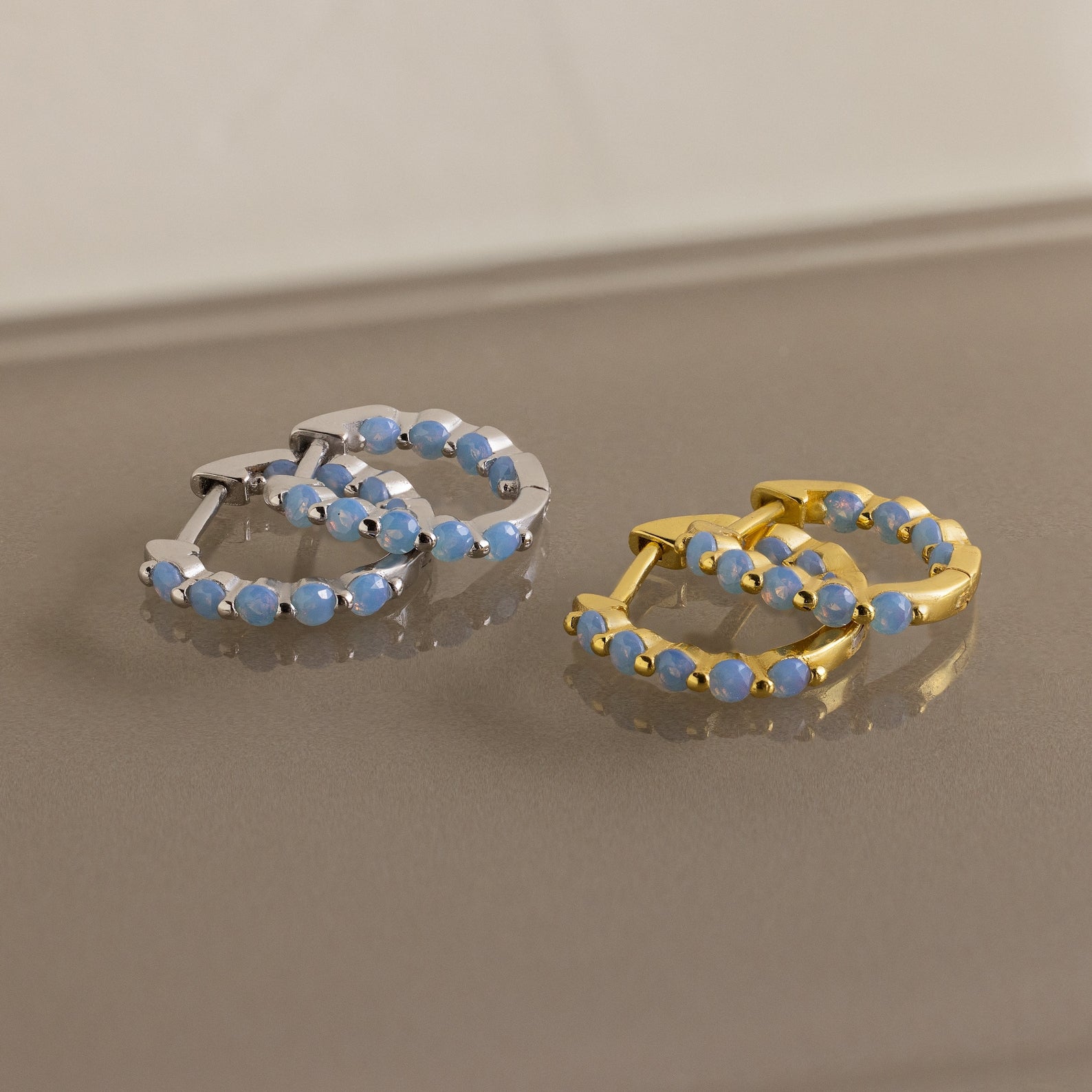 Two pairs of Inside Out Ivy Huggies—one in silver, one in gold—each set with delicate light blue gemstones, are elegantly displayed on a reflective surface.