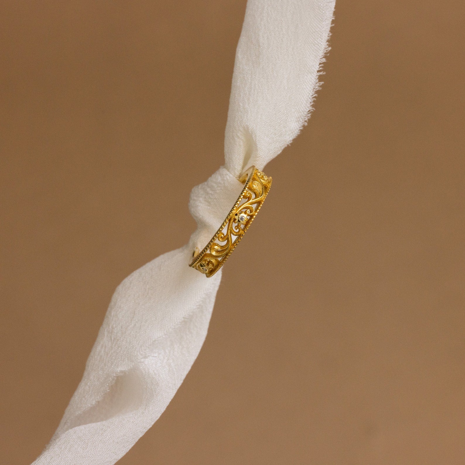The Filigree Flower Ring, a vintage-inspired gold band engraved with delicate floral details, is displayed on a white cloth with a beige background.