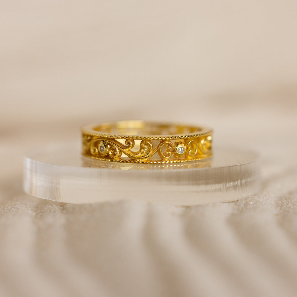 The Filigree Flower Ring features a gold band with intricate scrollwork and small embedded diamonds, evoking vintage charm and displayed elegantly on a clear, curved stand.
