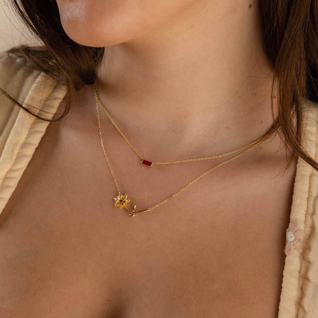 Woman wearing two gold necklaces: a Flower Birthstone Necklace with a red rectangular pendant and a personalized charm featuring a small leaf and berry.