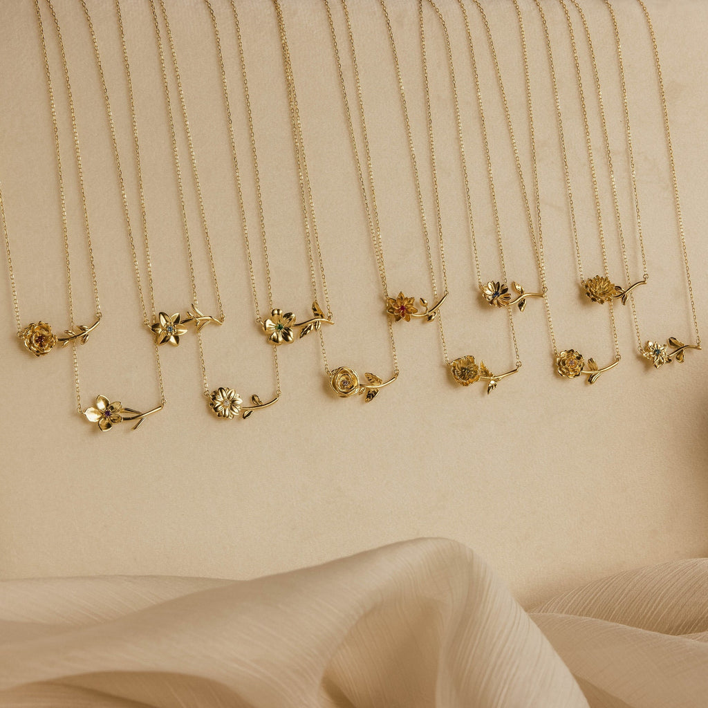 The Flower Birthstone Necklace features a gold flower pendant on a delicate chain, ideal as a personalized charm or custom necklace, beautifully displayed on a beige surface with soft fabric in the foreground.