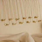 The Flower Birthstone Necklace features a gold flower pendant on a delicate chain, ideal as a personalized charm or custom necklace, beautifully displayed on a beige surface with soft fabric in the foreground.