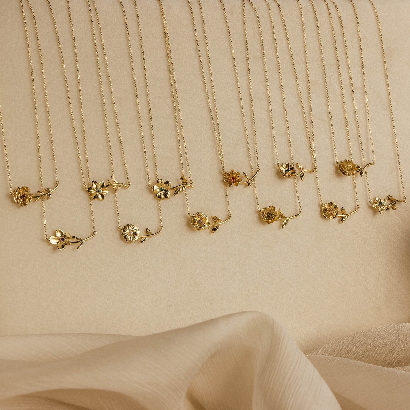 The Flower Birthstone Necklace features a gold flower pendant on a delicate chain, ideal as a personalized charm or custom necklace, beautifully displayed on a beige surface with soft fabric in the foreground.