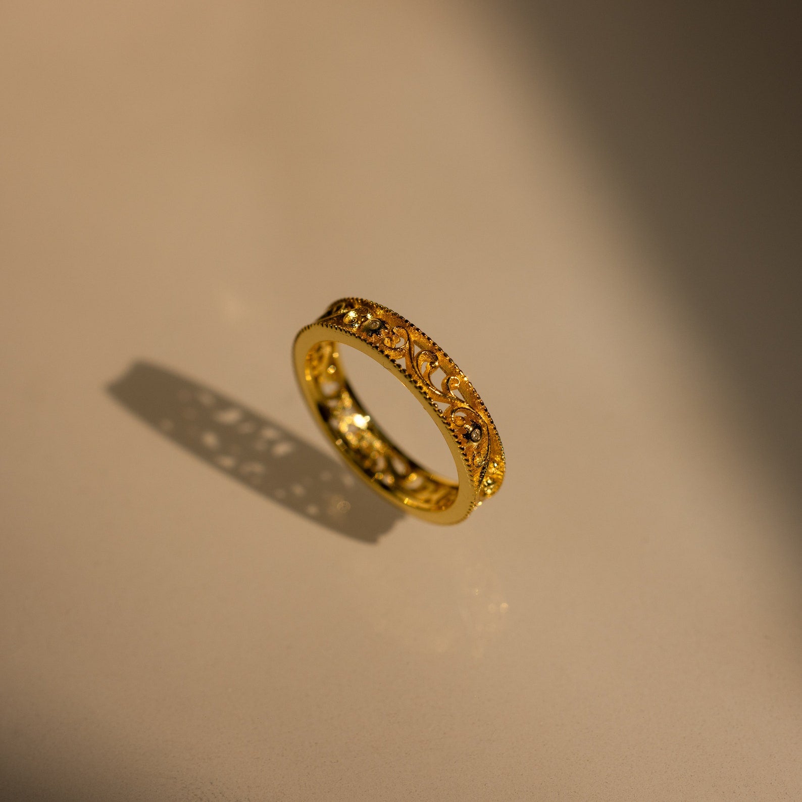The Filigree Flower Ring, featuring a vintage-inspired design and gold band, casts a delicate shadow on a smooth, light-colored surface.