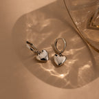 The Puff Heart Charm Huggies in Sterling Silver, shaped like hearts, rest on a beige surface with soft reflections from a glass object—ideal for anyone who loves elegant heart-shaped sterling silver earrings.