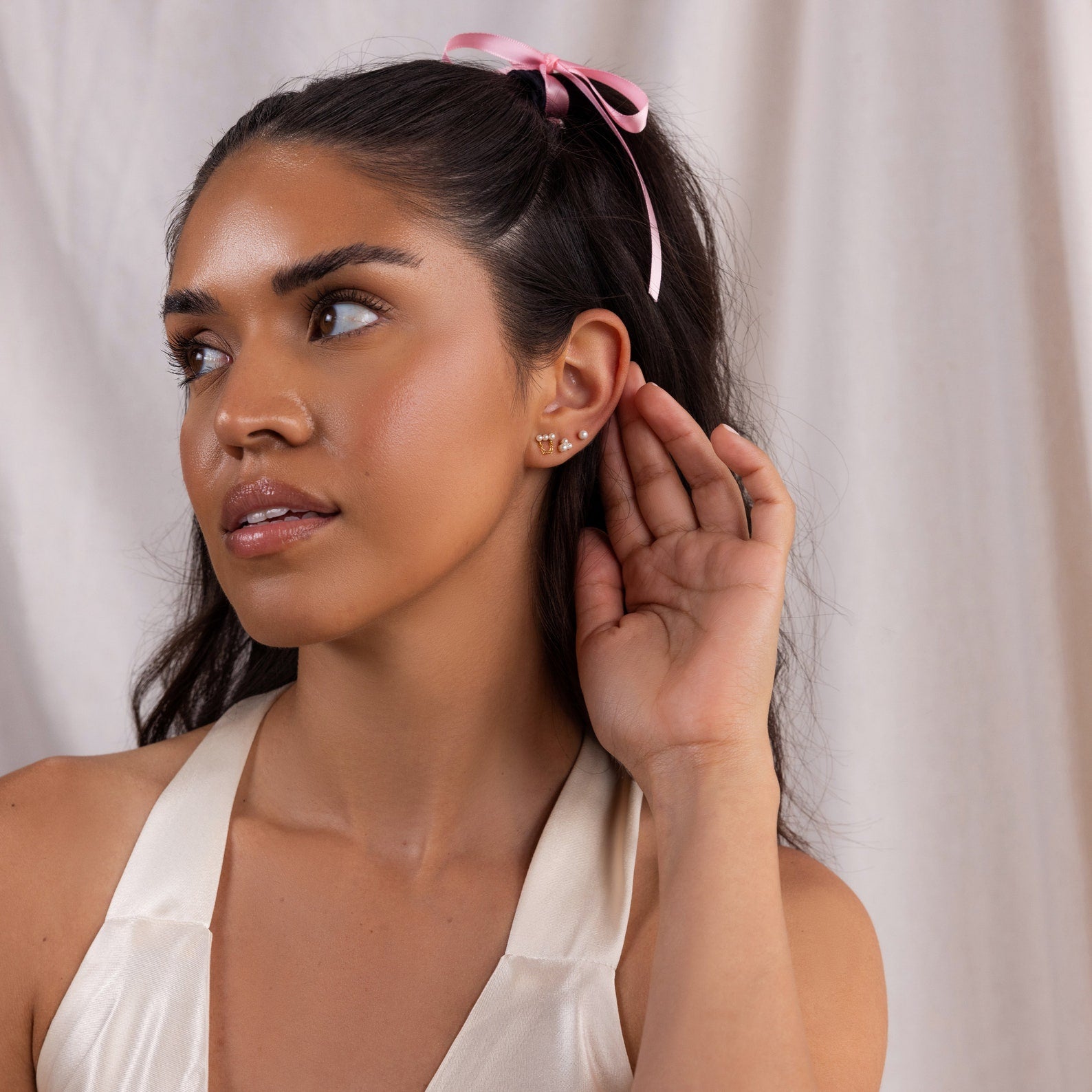 Woman with long dark hair, a pink ribbon, and elegant Trio Cluster Pearl Studs holds her hand to her ear, listening attentively.