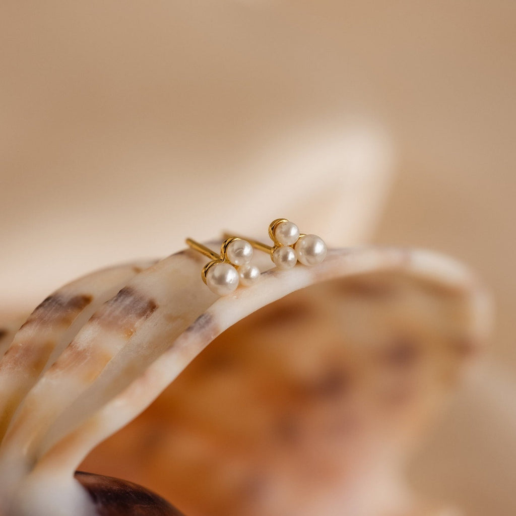 The Trio Cluster Pearl Studs, featuring gold pearls in delicate clusters, rest on a seashell against a soft beige background—ideal as bridal earrings or elegant wedding jewelry.