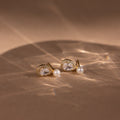 The Pearl Diamond Teardrop Studs feature gold settings with teardrop crystals and small pearls, displayed on a beige surface under soft lighting.