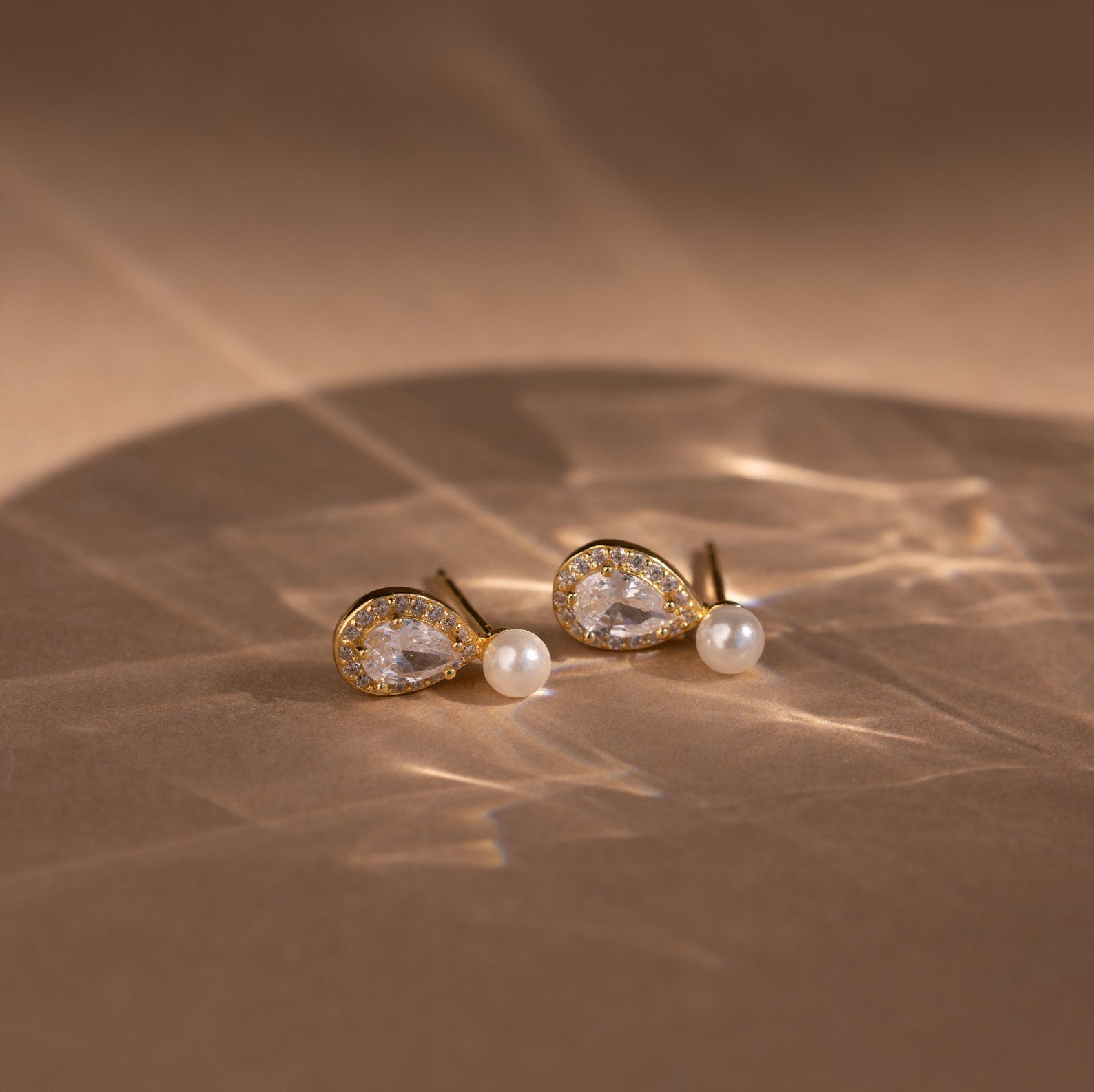 The Pearl Diamond Teardrop Studs feature gold settings with teardrop crystals and small pearls, displayed on a beige surface under soft lighting.