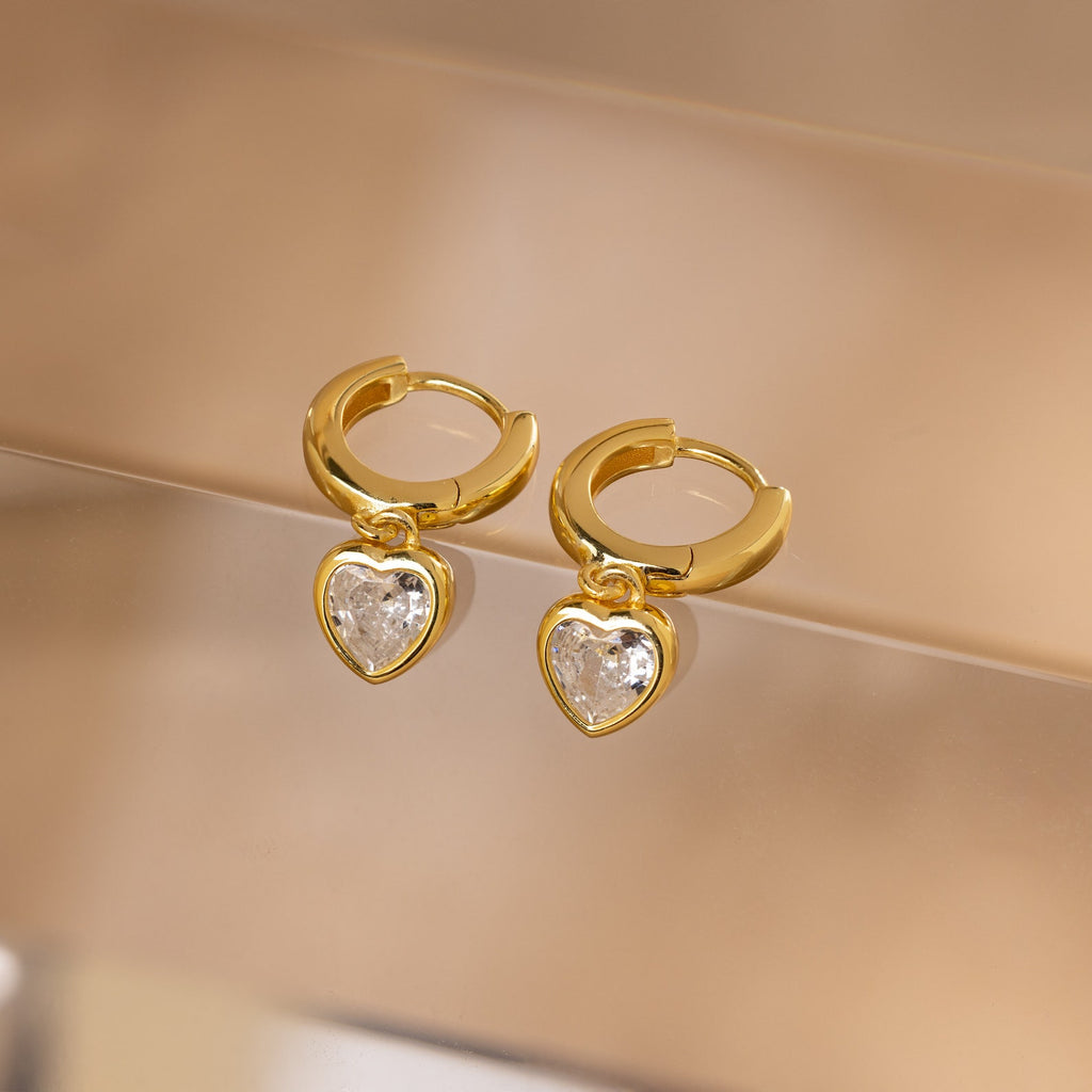 Diamond Heart Drop Huggies—coquette-inspired gold hoop earrings with sparkling diamond heart charms—displayed on a soft beige surface.