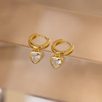 Diamond Heart Drop Huggies—coquette-inspired gold hoop earrings with sparkling diamond heart charms—displayed on a soft beige surface.