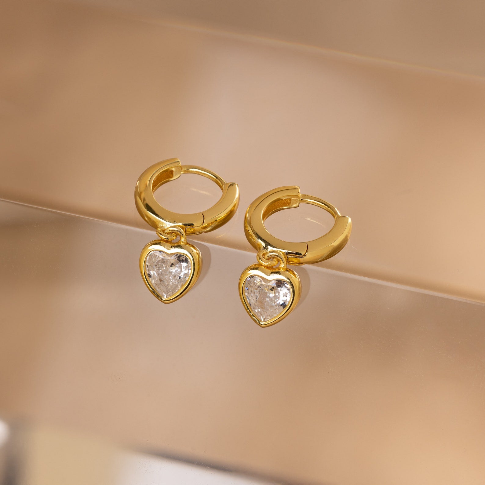 Diamond Heart Drop Huggies—coquette-inspired gold hoop earrings with sparkling diamond heart charms—displayed on a soft beige surface.