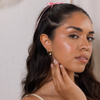 Woman with long dark hair, a pink ribbon, and gold earrings touches her face and looks at the camera, exuding coquette-inspired elegance with her Diamond Heart Drop Huggies sparkling subtly.