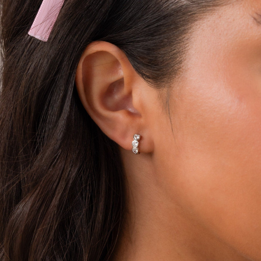 Close-up of a woman's ear wearing Diamond Bezel Heart Huggies—small silver earrings featuring three sparkling gemstones.