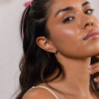 A woman with wavy hair and a pink headband wears Diamond Bezel Heart Huggies, delicately touching her chin.