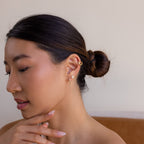 A woman with a low bun and multiple gold earrings gazes down thoughtfully, her hand near her chin, as if admiring new finds from the Mystery Earrings Box.