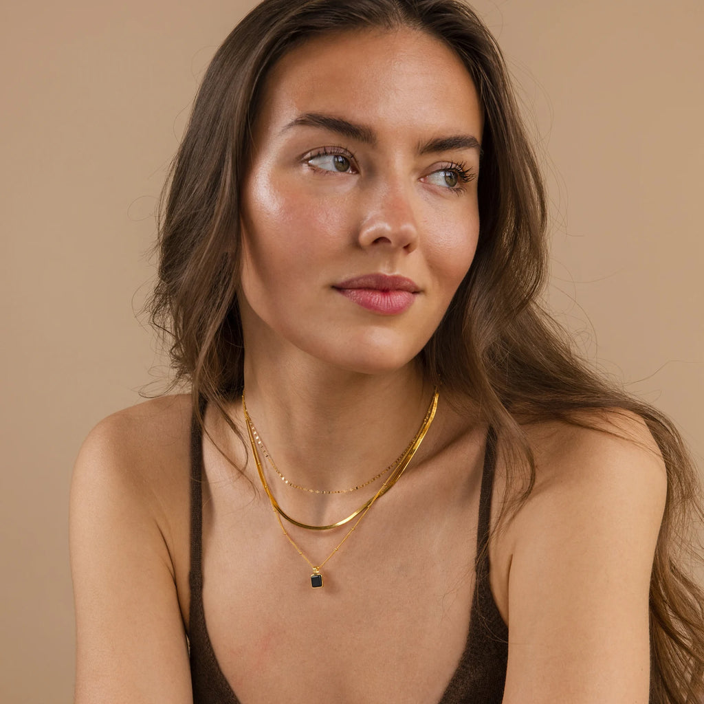 Woman with long brown hair wearing gold necklaces from our Mystery Necklace Box and a brown tank top, looking to the side.