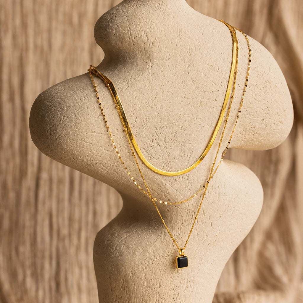 The Mystery Necklace Box features layered gold necklaces with unique chain styles and a black pendant, elegantly displayed on a neutral bust—perfect for enhancing any jewelry collection.