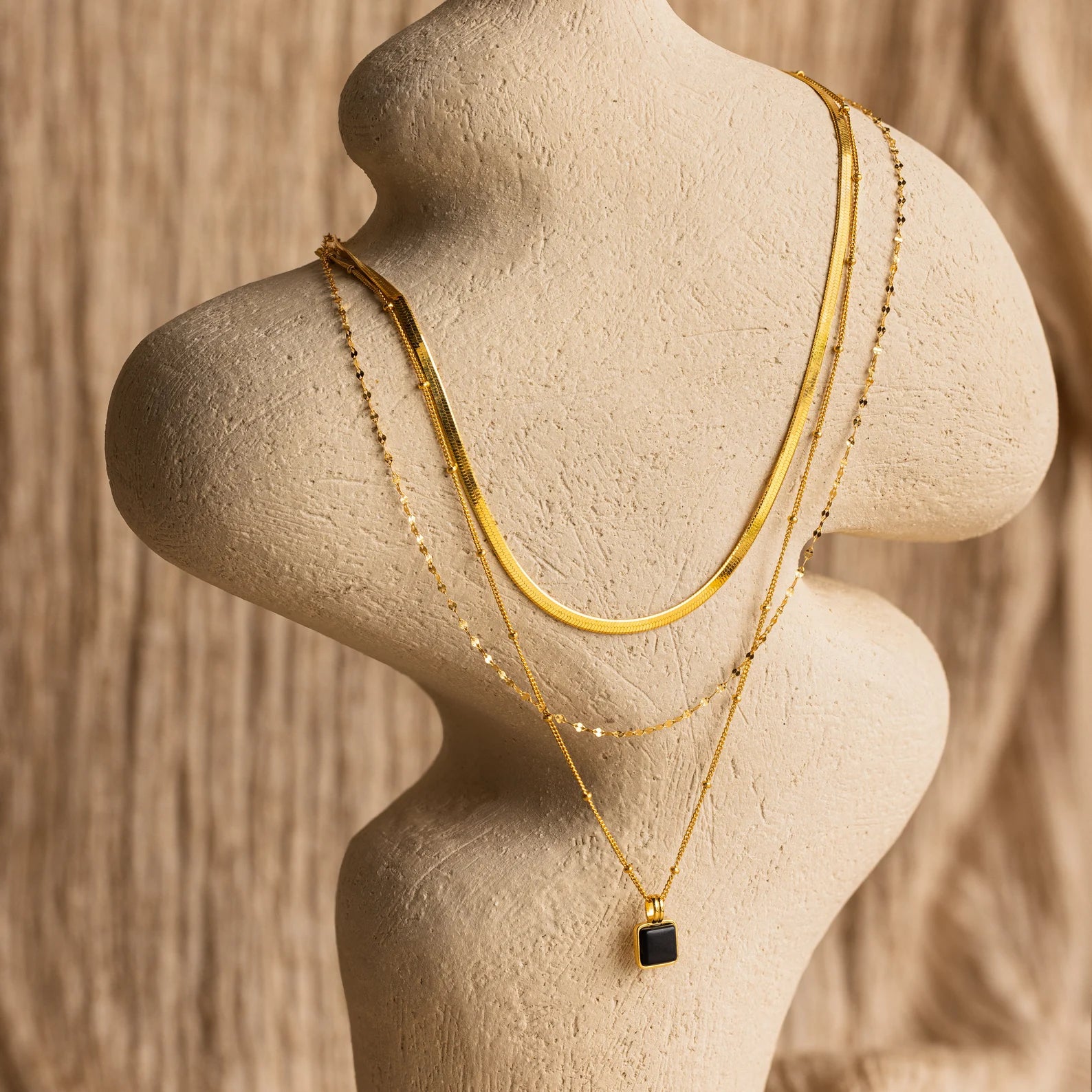The Mystery Necklace Box features layered gold necklaces with unique chain styles and a black pendant, elegantly displayed on a neutral bust—perfect for enhancing any jewelry collection.
