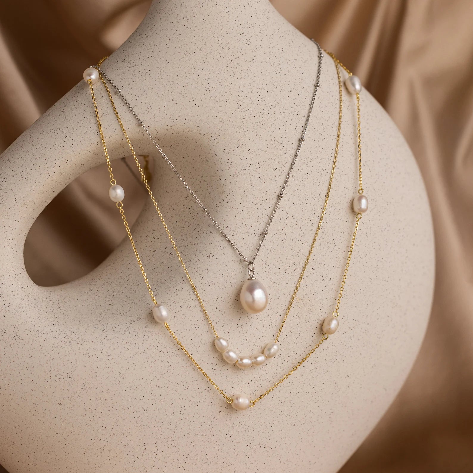 The Mystery Necklace Box from our jewelry collection features three layered pearl necklaces displayed on a smooth, curved beige mannequin against a satin background.