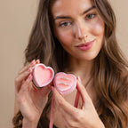 A woman smiles at the camera in a charming coquette style while holding the Pink Velvet Heart Ring Box, open to reveal two rings inside.