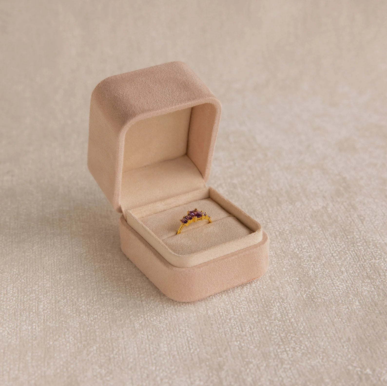 The Pink Suede Ring Box opens to reveal a gold ring with red gemstones, displayed on a neutral textured surface and highlighting its minimalist design.
